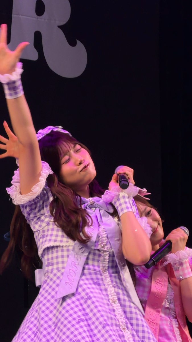 A young woman with long hair performing on stage, eyes closed and one arm raised, wearing a purple and white gingham dress with ruffled accents and holding a microphone.