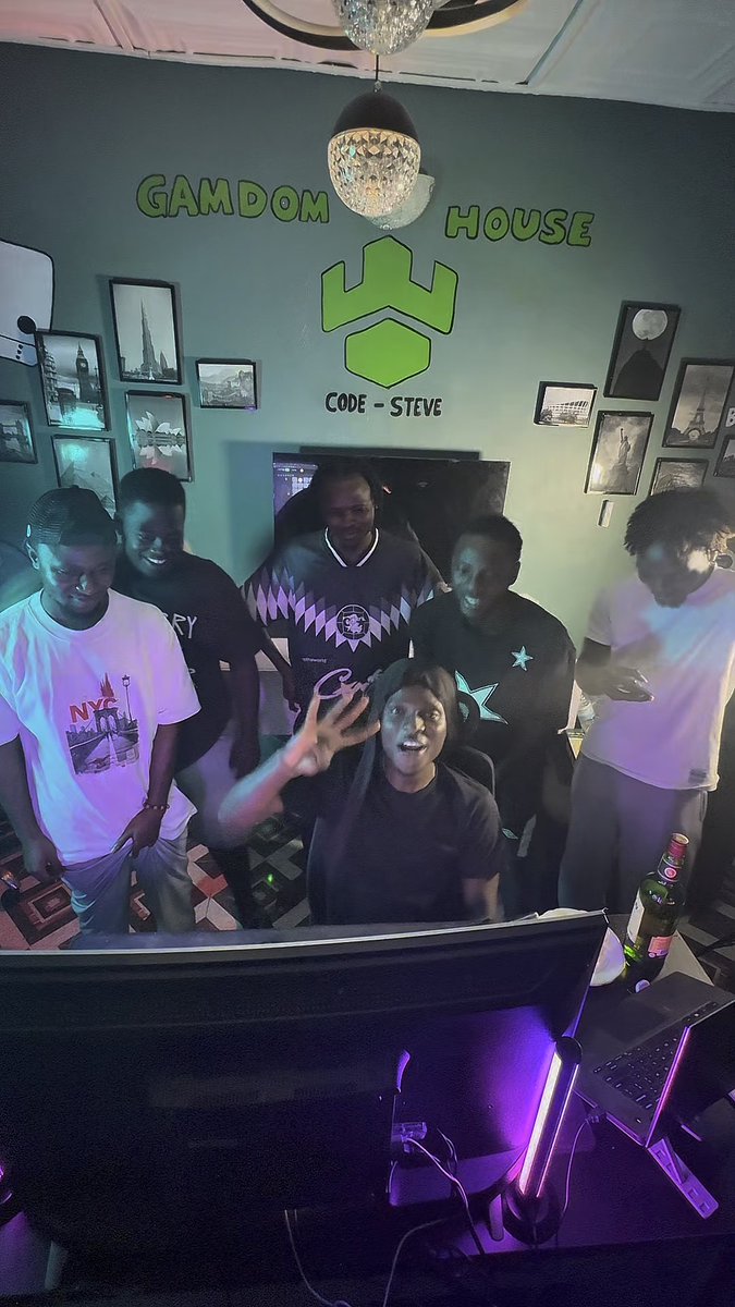 A group of six young people posing in a room with green walls, a 'GAMDOM HOUSE' sign, a 'CODE - STEVE' logo, and framed pictures, with one person in the center holding up a peace sign while sitting behind a computer monitor.