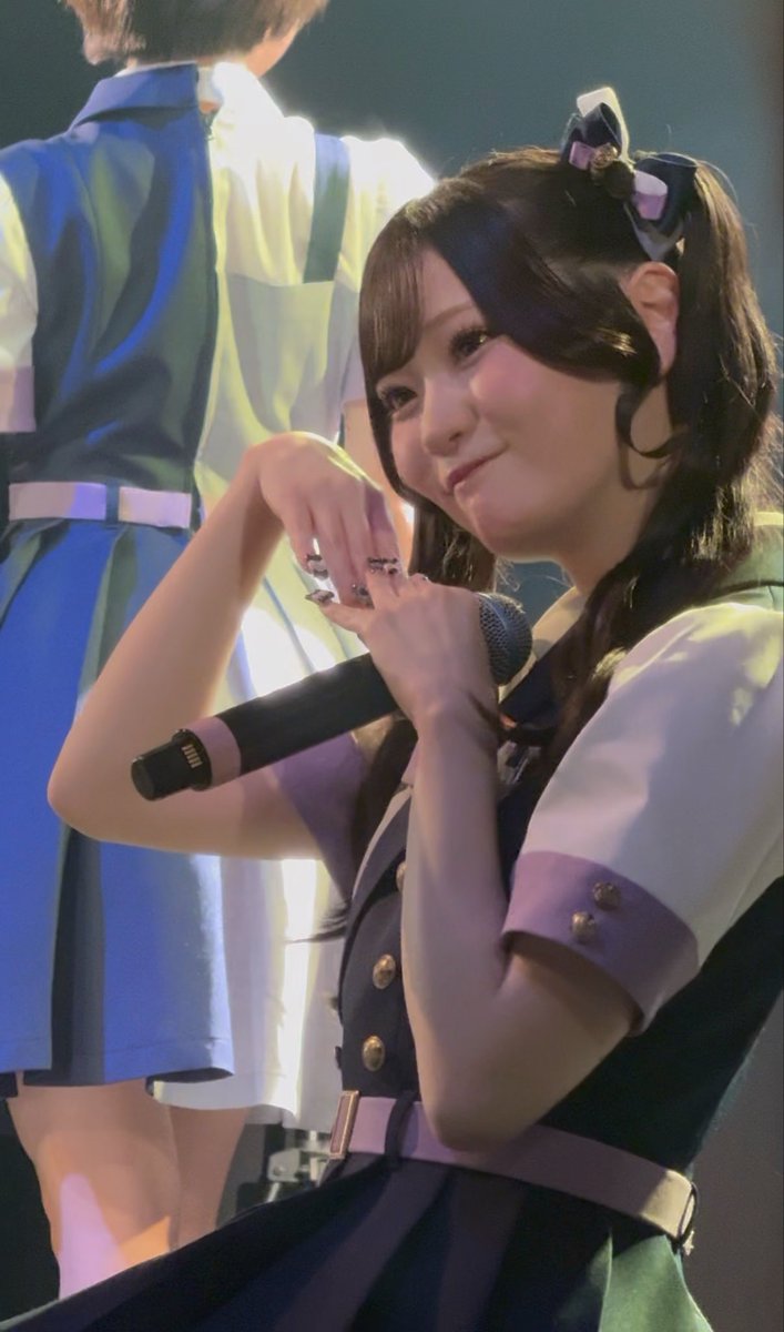 A young female performer with long dark hair styled in pigtails with ribbons holds a microphone on stage, wearing a navy and white uniform with gold buttons, as she smiles softly with her face turned to the side—showcasing the adorable side profile referenced in the caption.