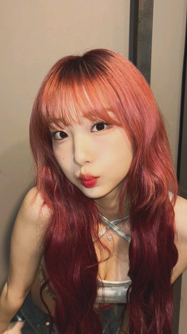 A selfie of a young woman with long, wavy red hair, blunt bangs, and red lipstick, wearing a silver strappy top and layered necklaces.