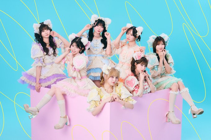 Seven members of a Japanese idol group pose in pastel-colored frilly outfits and fluffy bear-ear headbands against a turquoise background with pink geometric shapes, smiling and making cute gestures. The image accompanies a post announcing the launch of "UP-T TALK," a new event offering 1-minute video calls with fans starting at 8 PM, with the URL talk.up-t.jp/universal-link provided for app download.
