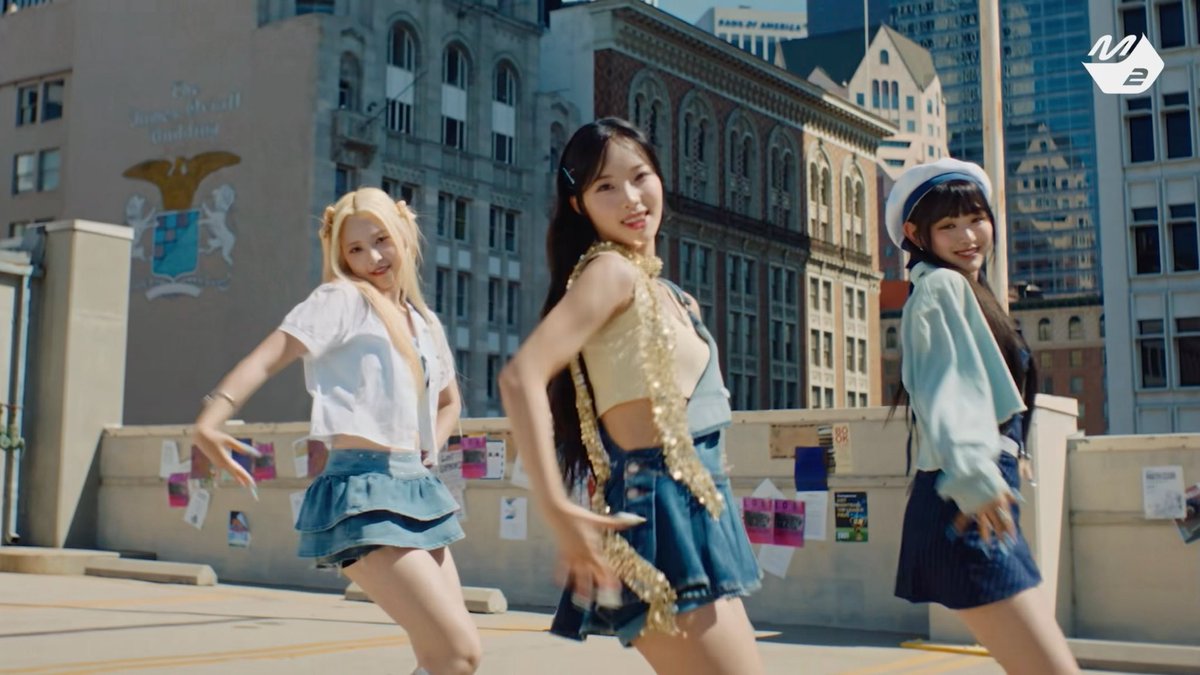 Three women dancing on a rooftop in a city setting, wearing stylish outfits including skirts and a sailor hat, with a 'NB' logo in the corner.