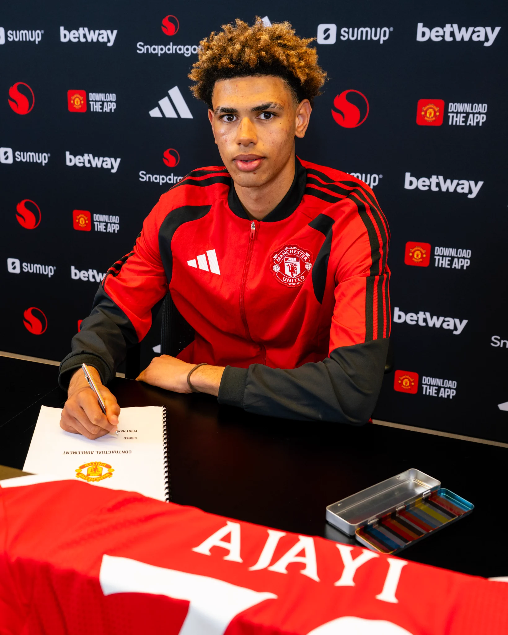 Noah Ajayi wearing a red and black United jacket signing his contract in front of a sponsored backdrop.
