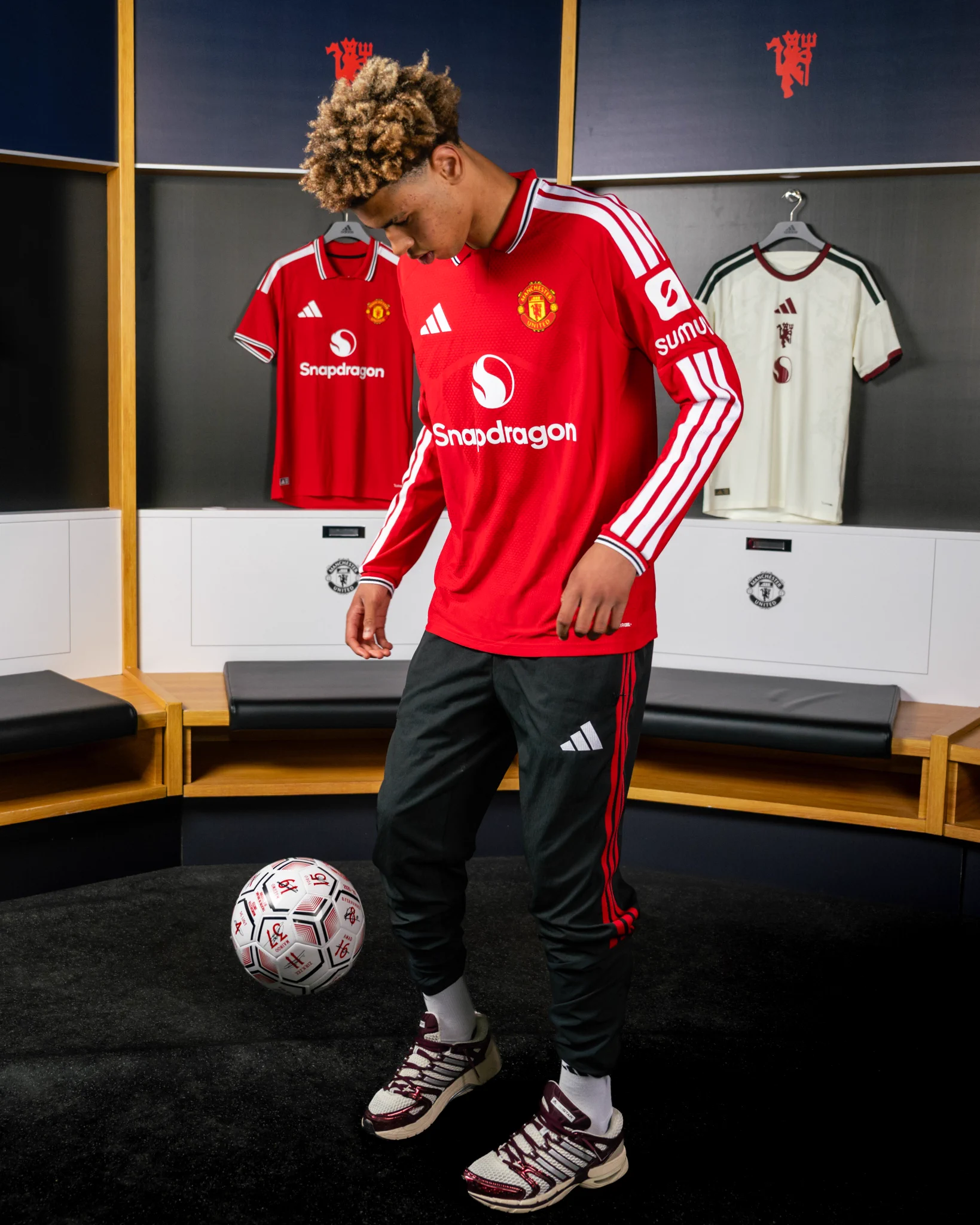Noah Ajayi wearing a long sleeve 2026/27 United home shirt and black tracksuit pants doing keepy uppies in the dressing room.