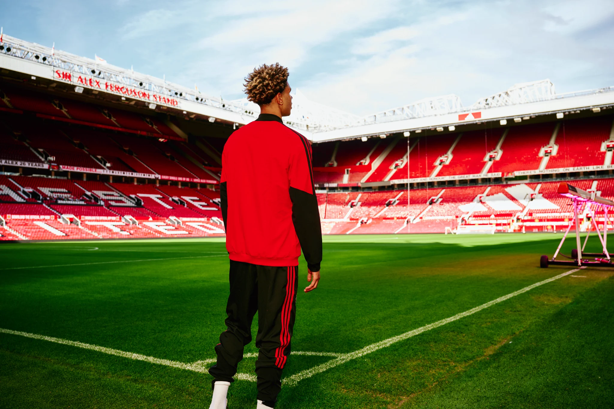 Noah Ajayi looks out at an empty Old Trafford beneath blue skies from pitchside while wearing a red and black tracksuit.