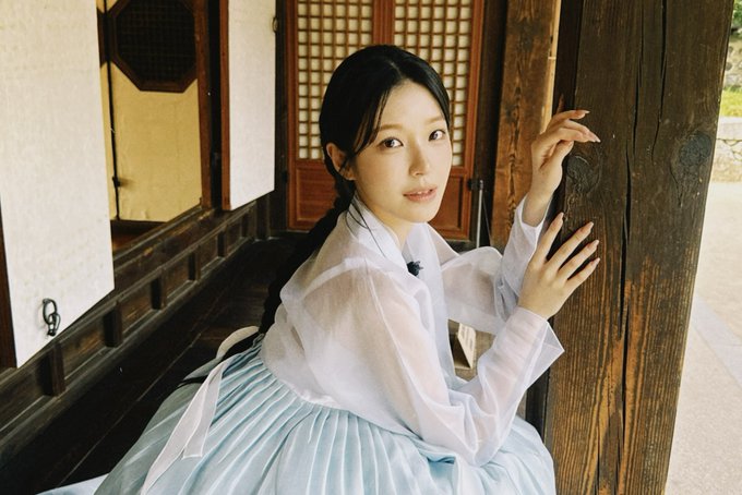 A young woman wearing a white sheer blouse and light blue traditional Korean hanbok skirt leans against a dark wooden pillar in front of a traditional Korean building with lattice doors.