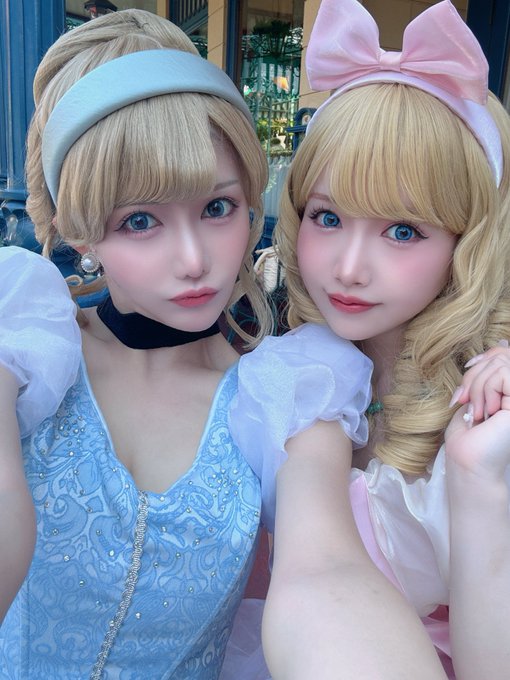 Two women in elaborate Disney princess cosplay, one dressed as Cinderella in blue and the other in a pink bow and dress, pose together for a selfie.