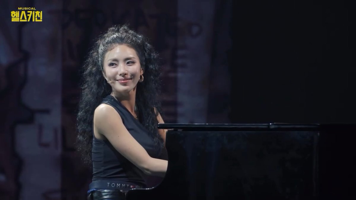 A woman with long curly hair, wearing a dark sleeveless top with 'TOMMY' visible on the waistband, sits at a piano during a musical performance, smiling at the camera with the Korean text 'MUSICAL 엠스기전' displayed in the upper left.