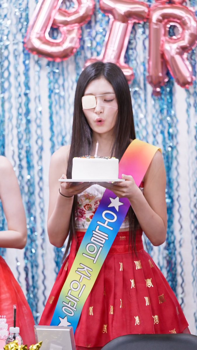 A young woman with long dark hair wearing an eye patch, a floral top, and a red skirt blows out a candle on a birthday cake while wearing a rainbow sash that reads 'You can't [do]' at what appears to be a birthday celebration with pink letter balloons spelling 'STAR' in the background.