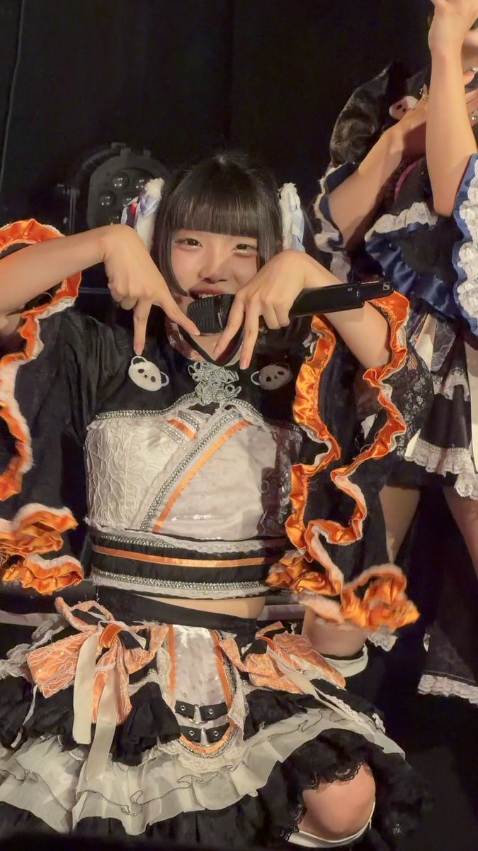 A young performer in an elaborate black, white, and orange ruffled costume holds a microphone and makes a peace sign gesture, with another performer partially visible behind her.