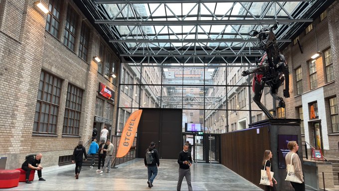 A photograph of an indoor industrial-style venue with brick walls, a glass ceiling, and a large dark horse statue on a pedestal, where several people are walking around an 'ETCHEL' banner.