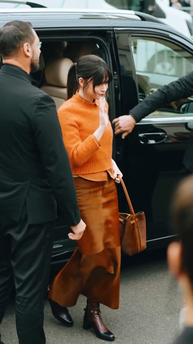 A woman with dark hair tied back, wearing an orange sweater and brown velvet skirt, exits a black vehicle while being assisted by men in black suits.