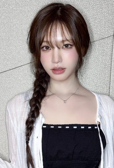 A portrait of a young woman with a long side braid, wearing a white top over a black top, a small heart necklace, and pink lipstick, posed against a stone wall.