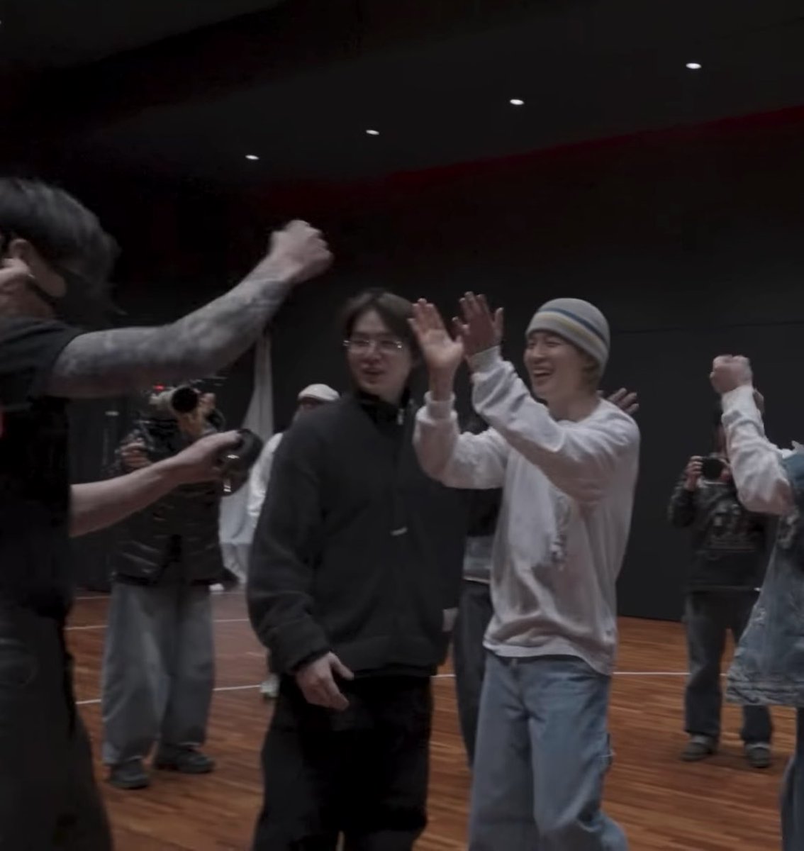 Two young men in a studio setting smiling and high-fiving, with one wearing a beanie and white shirt, while a cameraman films them and others celebrate in the background.
