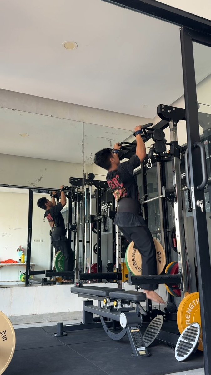 A person in a black t-shirt and dark pants performs a pull-up exercise on a cable machine in a gym, with their reflection visible in a large mirror behind them.