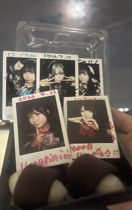 A hand holds up a clear plastic case displaying four polaroid-style photos of a young woman making various hand gestures, with Japanese text and dates like "2026.7.17" and "2026.9.22" handwritten in red marker on the white borders.