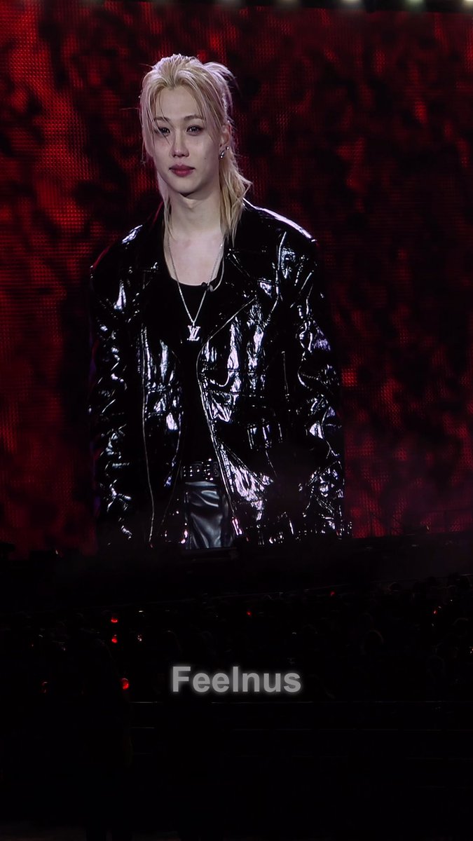 A blonde performer wearing a black patent leather jacket and an LV pendant stands on a stage against a red backdrop, with "Feelnus" visible at the bottom of the screen.