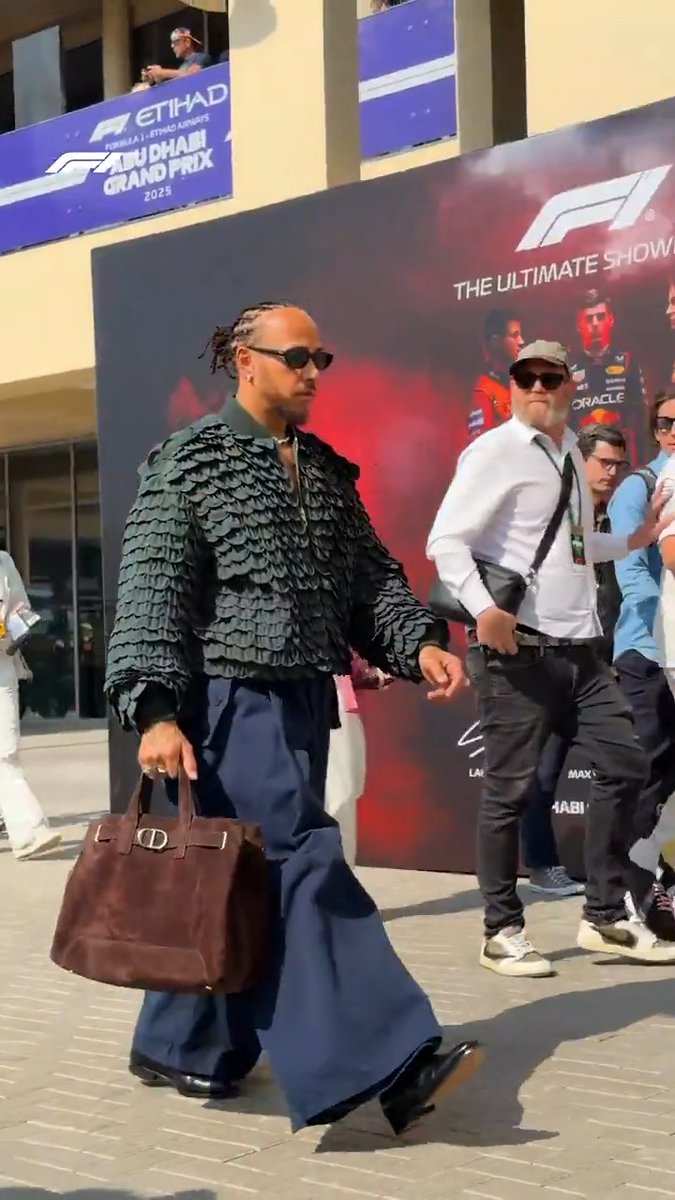 Lewis Hamilton makes a bold fashion statement in a textured black jacket and wide-leg trousers while carrying a brown suede bag outside the Formula 1 paddock at the 2025 Etihad Abu Dhabi Grand Prix, with "F1 THE ULTIMATE SHOWDOWN" visible on the backdrop behind him.