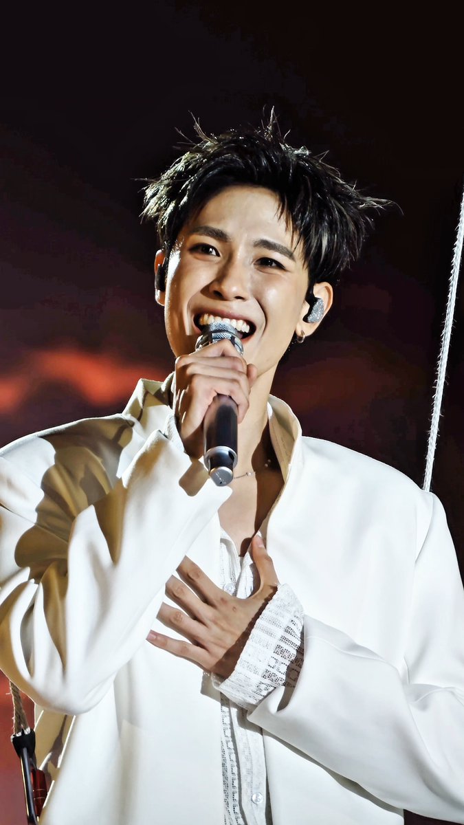 A young male performer with messy dark hair smiles broadly while singing into a rhinestone-encrusted microphone, wearing a white shirt with lace cuffs during a live concert performance.