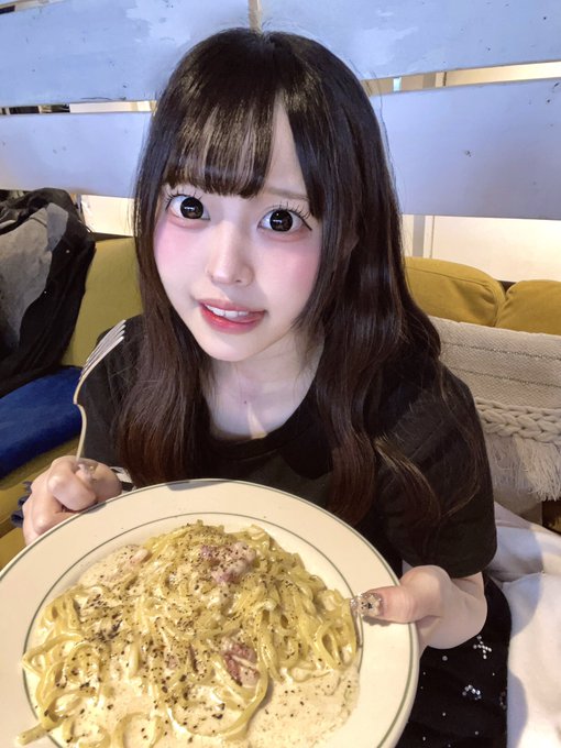 A young woman with long dark hair and striking makeup smiles while holding a large plate of creamy carbonara pasta in front of a couch.