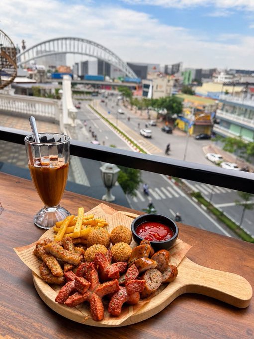 A wooden board laden with assorted foods including fries, onion rings, sausage, and fried items served with ketchup, accompanied by a glass of iced coffee, set on a balcony overlooking a city street with a large arched bridge in the background.