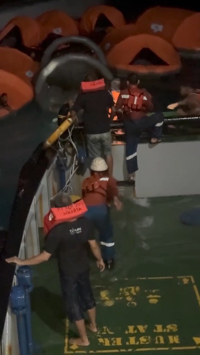 A coal ship rescues passengers of the KM. Virgo at night, with crew members wearing orange "Pertamina Jakarta" life jackets and hard hats assisting people from an inflatable lifeboat onto the deck of the vessel amid rough seas. The image captures the chaotic scene of the rescue operation, illustrating the difficult conditions mentioned in the post about the terrifying high waves and darkness.