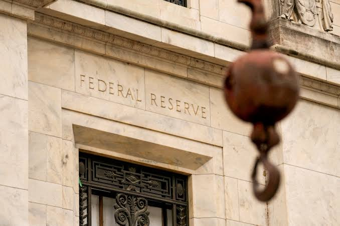 The Federal Reserve building is shown with its name engraved in stone above an ornate doorway, while a copper-colored pendant light hangs in the foreground. The image visually reinforces the article's focus on the Fed's potential regulatory changes, which could ease oversight thresholds and unlock a new wave of bank mergers and acquisitions.
