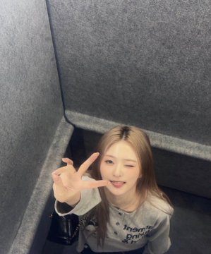 A smiling woman with long brown hair poses with a peace sign while wearing a gray "Indigo" t-shirt in an elevator. The accompanying text explains the textured gray walls are protective covers, with the photo likely captured through a rear camera reflection causing the flipped orientation.