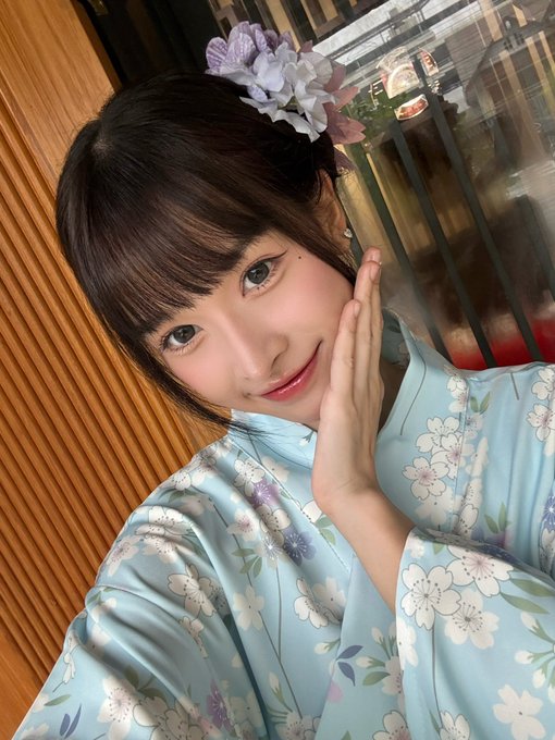 A young woman smiles for a selfie while wearing a light blue floral kimono and a matching purple and white hydrangea hair accessory, posing near a wooden paneled wall during a Japanese matcha workshop at NOAN Japanese Matcha & Dining in the Ekkamai branch, as the caption mentions receiving the cute hair tie as a souvenir from the event.