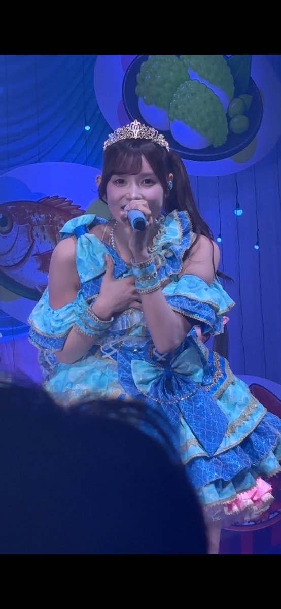 A performer in an elaborate blue ruffled idol dress with a tiara sings into a microphone on a stage with blue lighting and dessert-themed decorations.