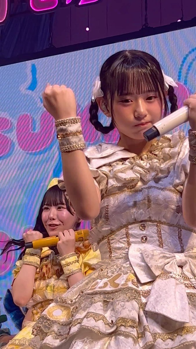 Two young performers in elaborate frilly white-and-gold and yellow costumes strike energetic poses with microphones on a colorful stage, with the girl in front raising her fists and the one behind smiling brightly while holding her mic horizontally.