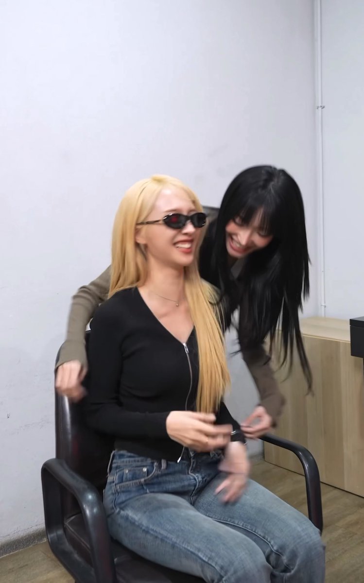 Two young women are smiling and laughing together in an indoor setting; one with long blonde hair wearing sunglasses and a black top sits in a chair, while another with long dark hair stands behind her.