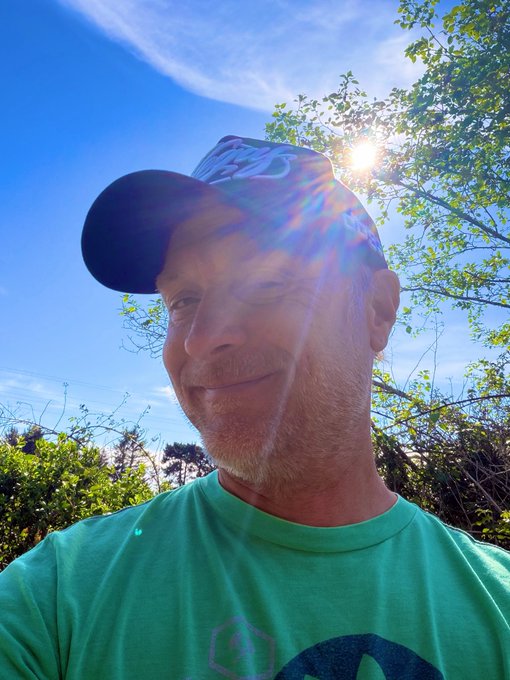 A selfie of a smiling older man with a gray beard wearing a black cap and green t-shirt, standing outdoors with the sun shining brightly behind him and trees in the background.