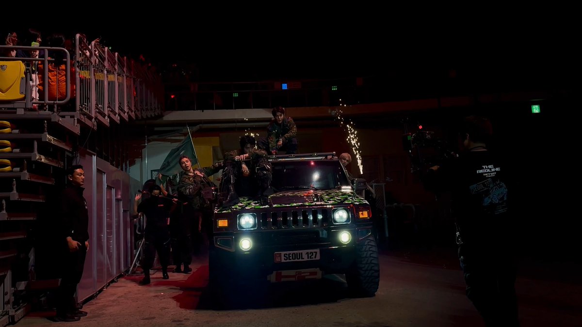 A Hummer with the license plate "SEOUL 127" drives through a dark venue with armed performers in military-style outfits on and around it, while crew members film the scene and spectators watch from an elevated platform. The image ties to NCT 127's "The Redline" tour, showcasing a cinematic concert moment that matches the dramatic, action-packed theme of their NEO CITY performances.