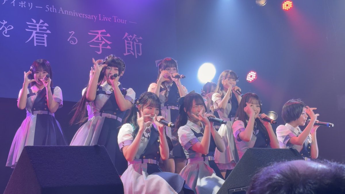 A group of Japanese idol performers in matching blue and white outfits sing and pose on stage at a live concert, with '5th Anniversary Live Tour' and Japanese text visible on the backdrop behind them.