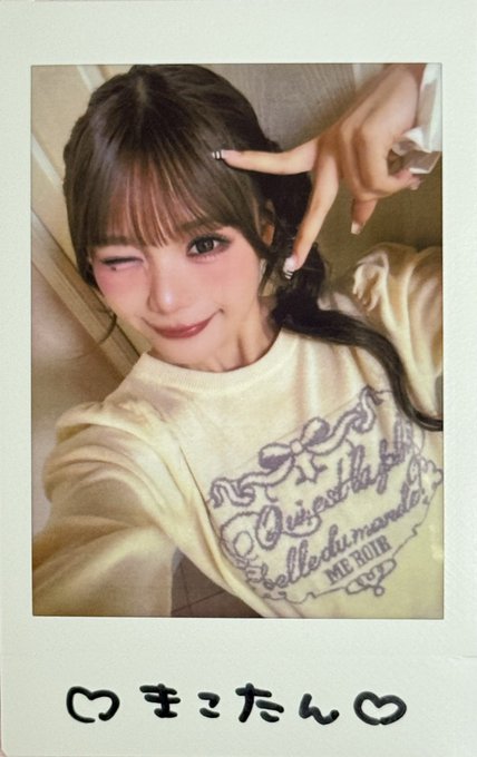 A Polaroid-style selfie of a young woman winking and making a peace sign, wearing a cream sweater with purple designs; the bottom border has handwritten Japanese text 'つまこたん♡'.