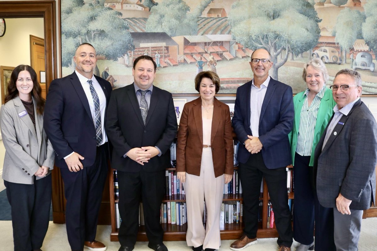 A group of seven adults pose for a photograph in an office or meeting room. They are standing in front of bookshelves and a large mural depicting a rural village and fields. Some people wear name badges; no prominent text is visible.