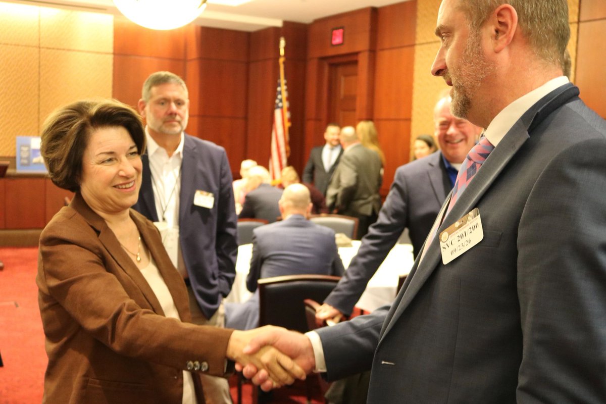 A woman in a brown blazer shakes hands with a man in a suit at a formal indoor gathering. Several people stand and sit in the background, with an American flag and tables in the room. Name badges and other text are visible.