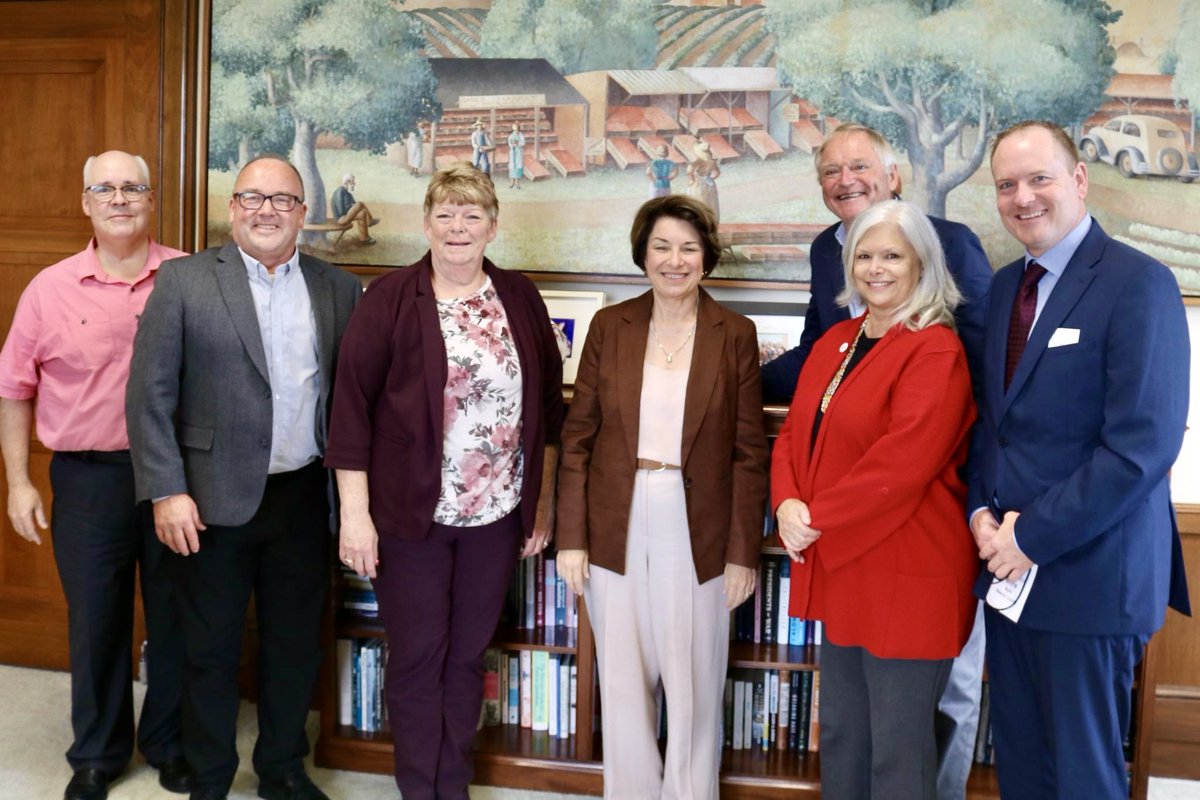 A group of seven adults pose for a photo indoors in front of a bookcase and a large mural. Some people hold papers; there is no prominent readable text.