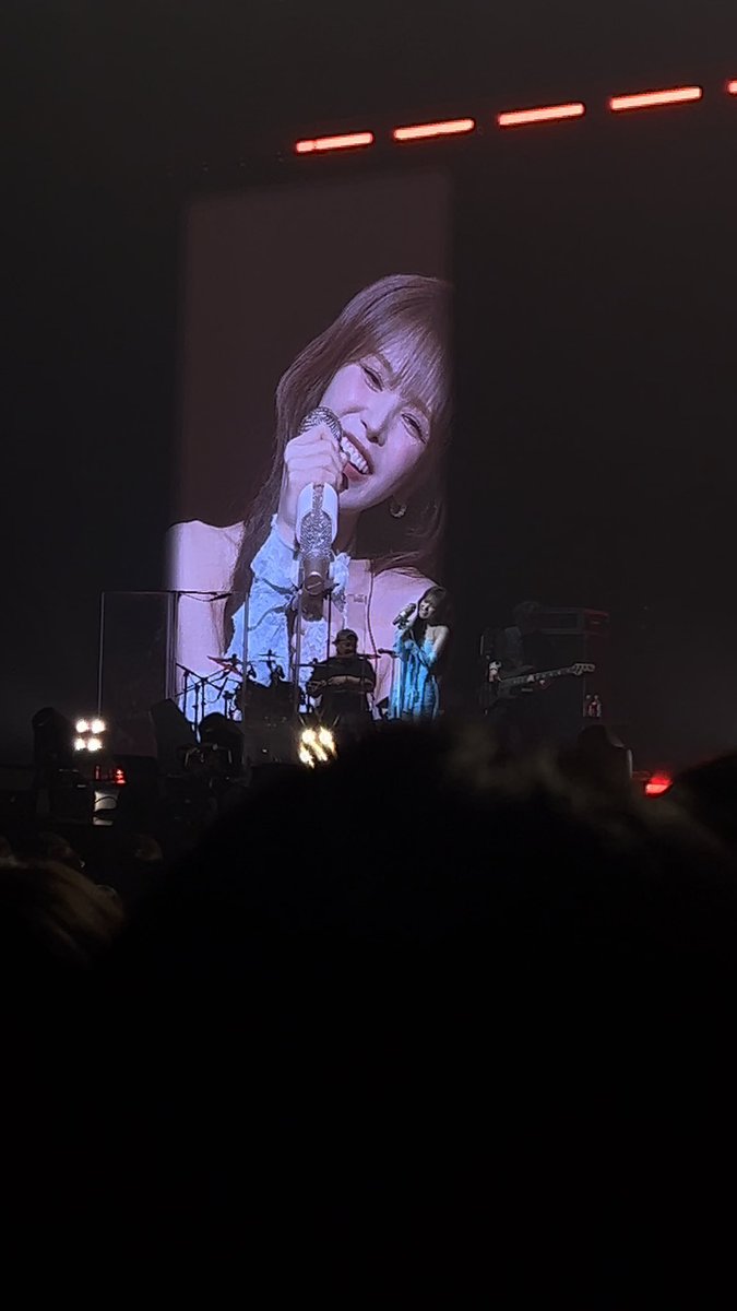 A concert view shows a female singer performing on stage while a large screen displays a smiling close-up of her face holding a sparkly microphone.