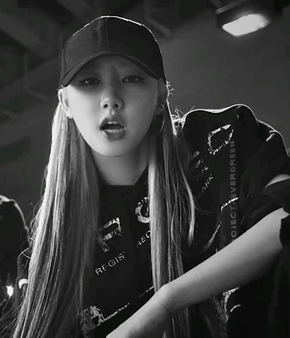 A black-and-white portrait of a young woman wearing a black cap and oversized graphic t-shirt, looking directly at the camera with her mouth slightly open.