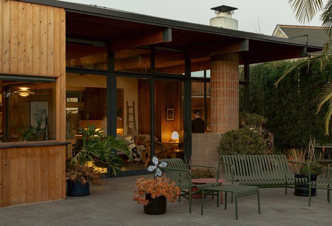 A warm, golden-lit midcentury modern home at dusk features a deep overhanging roof, large glass walls, and wood-clad columns opening onto a stone patio with green metal benches and potted plants, including a decorative flower sculpture. The visible interior—warm wood tones, vintage furnishings, and a person in a cap—evokes the lived-in character of a historic Black modernist residence.