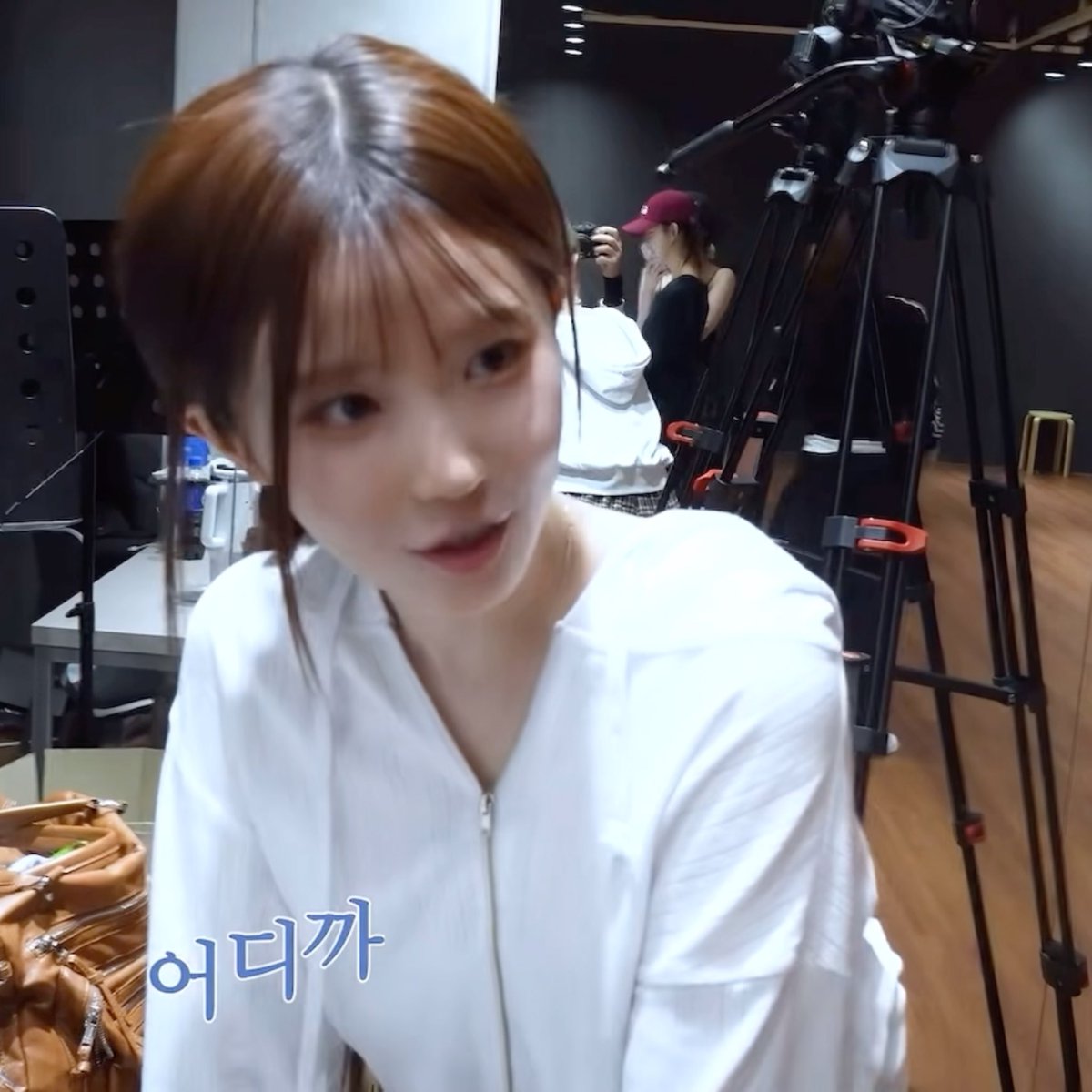 A young woman with short brown hair in a white zip-up jacket poses in what appears to be a photography studio, with Korean subtitle '어디까' (where to) visible.