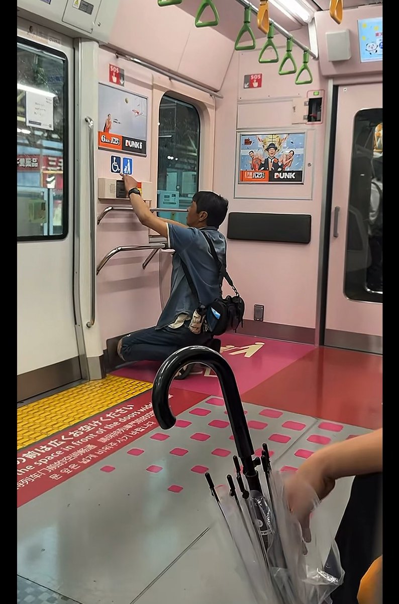Inside a pink train car decorated with anime posters, a man sits on a metal seat while reaching up to press a door control button, with a multi-jointed umbrella and another passenger visible in the foreground.