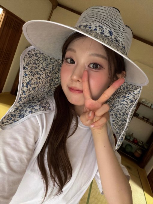 A young woman wearing a white t-shirt and a wide-brimmed floral sun hat poses for a selfie, making a peace sign with her fingers.