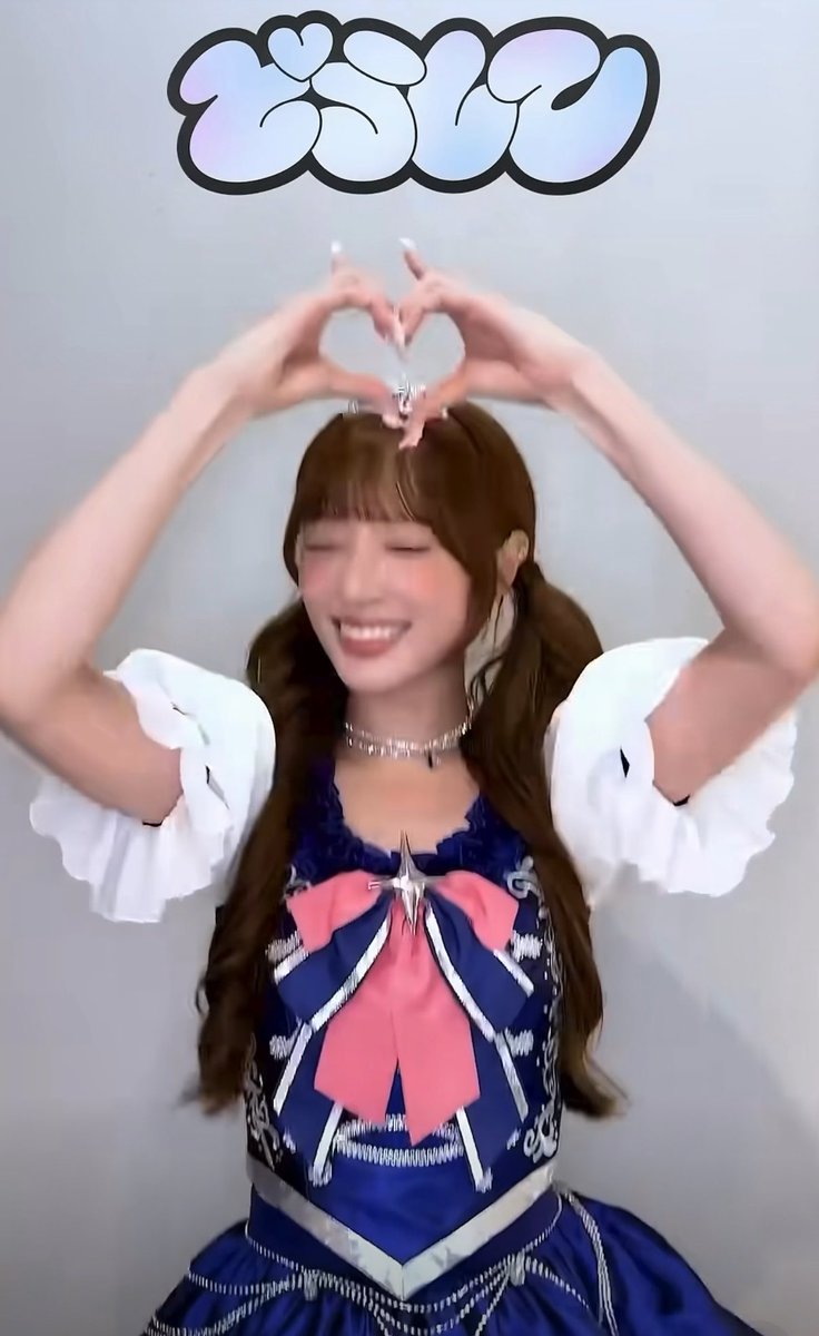 A young woman with brown hair in pigtails, wearing a blue and white dress with a large pink bow, smiles with her eyes closed while forming a heart shape above her head with her hands.