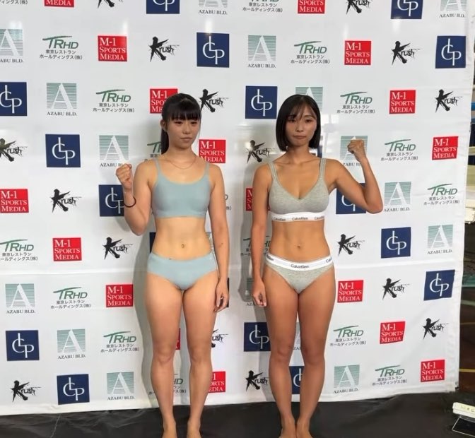 Two women pose in athletic underwear in front of a sponsor backdrop featuring logos for TRHD, AZABU BLD., Krush, M-1 SPORTS MEDIA, and CP.