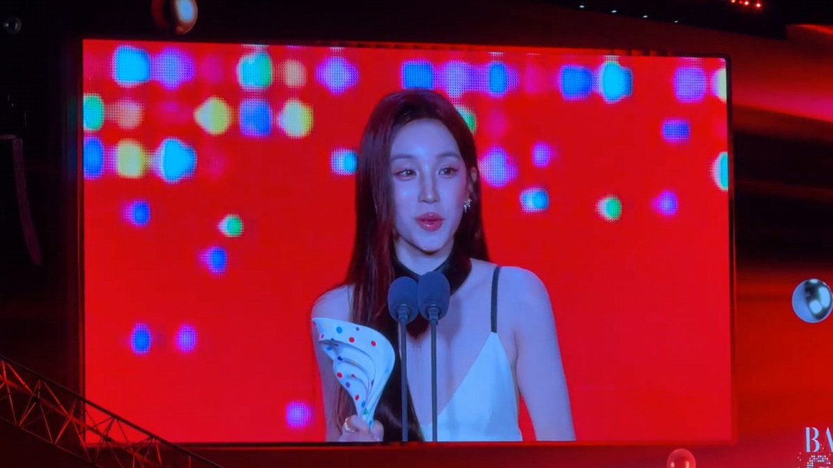 A woman speaks at a podium holding a trophy, displayed on a large screen with colorful bokeh lights against a red background.