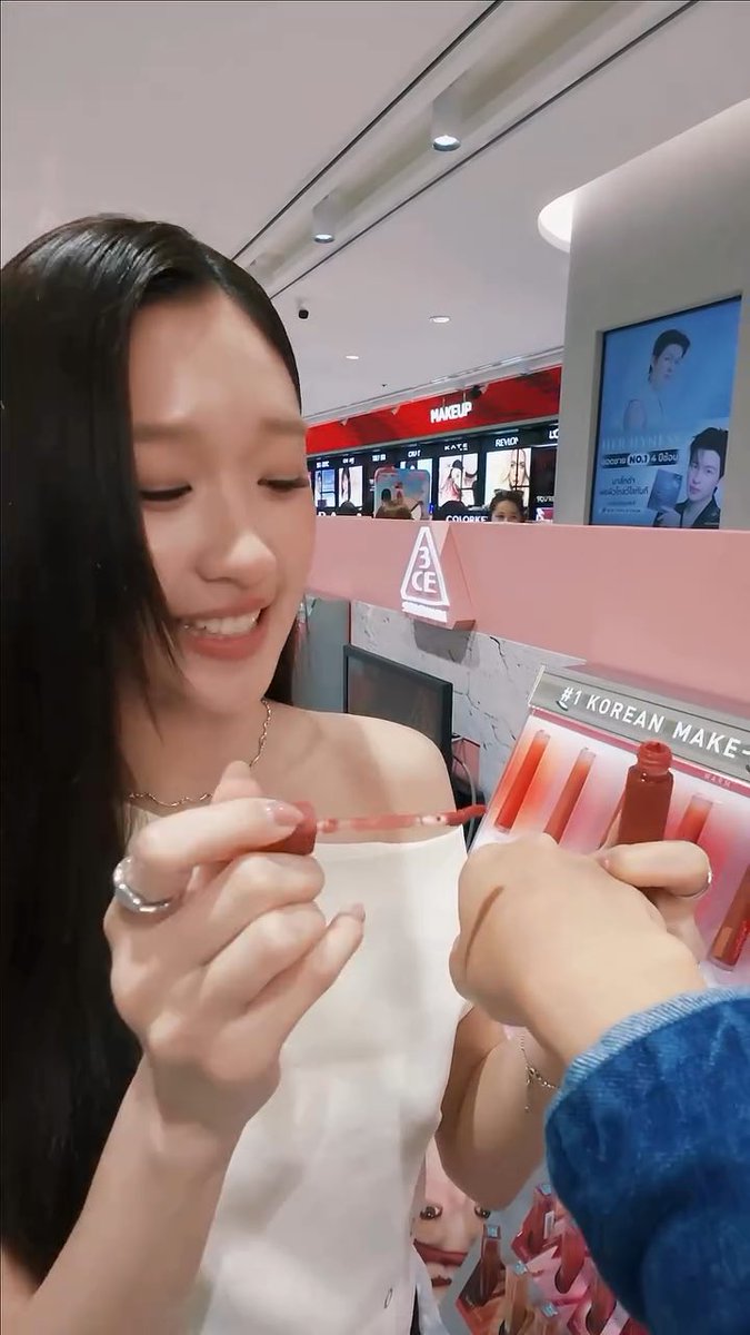 A woman smiling while holding up a lip gloss applicator in a cosmetics store, with "3CE" branding visible and text reading "#1 KOREAN MAKEUP" on the display.