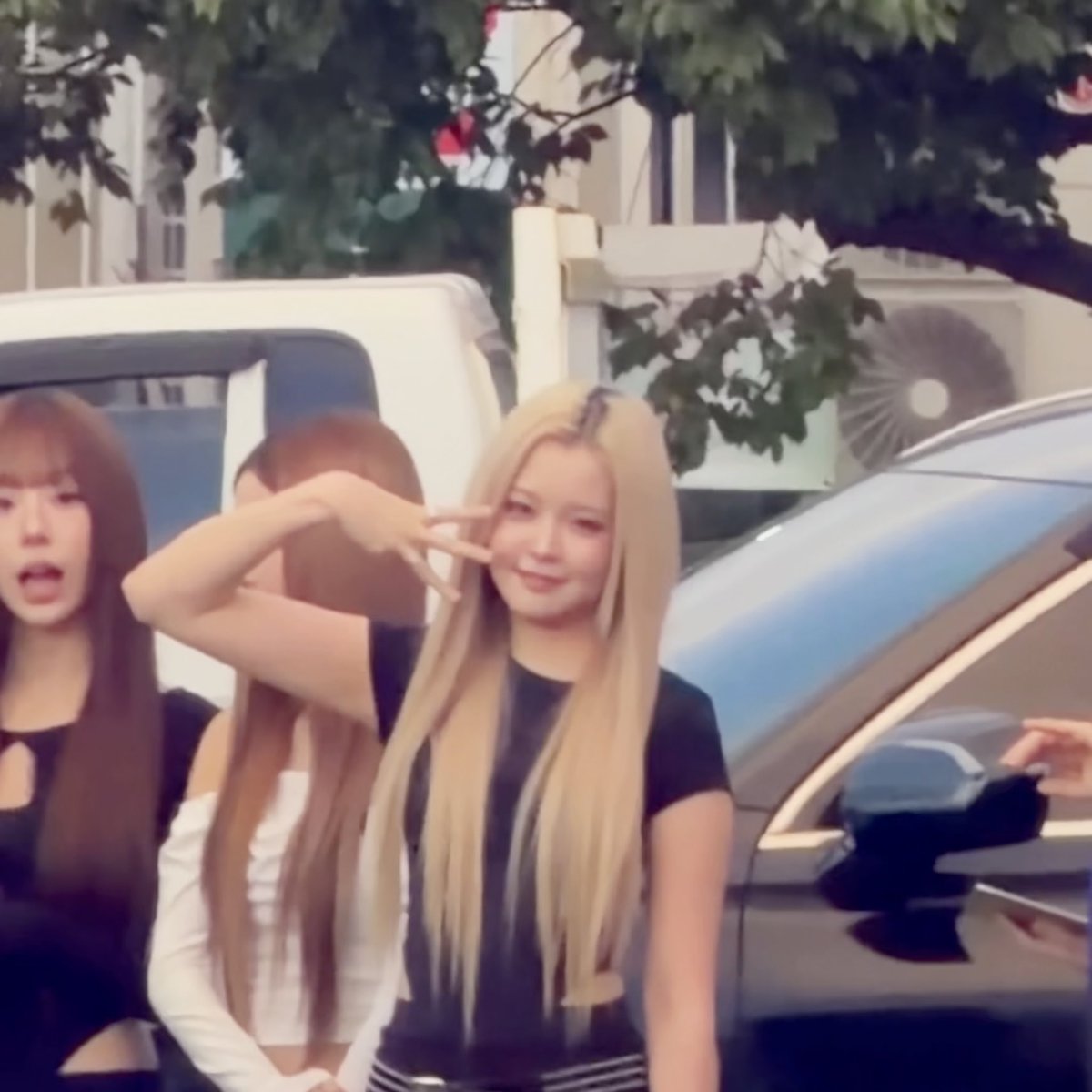A group of young women stand outside near a vehicle, with a blonde woman in the foreground making a peace sign while others stand behind her.